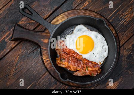 Bacon croccante e uovo fritto che sfrigolano in padella di ghisa su legno rustico, texture ricche ideali per temi di colazione e cucina gourmet. Foto Stock