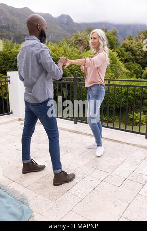 Diverse coppie senior che ballano e tengono le mani sulla terrazza piastrellata vicino alla ringhiera nera e alla coperta piegata Foto Stock
