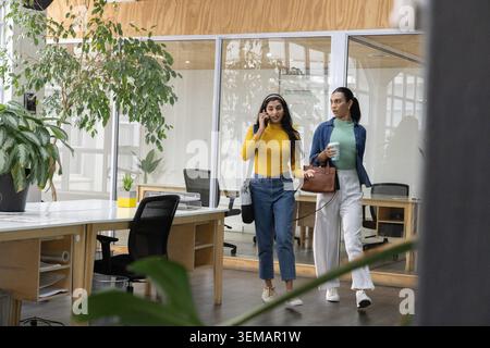 Diverse collaboratrici che camminano in ufficio passando davanti a banchi di legno, parlano al telefono e tengono un caffè Foto Stock