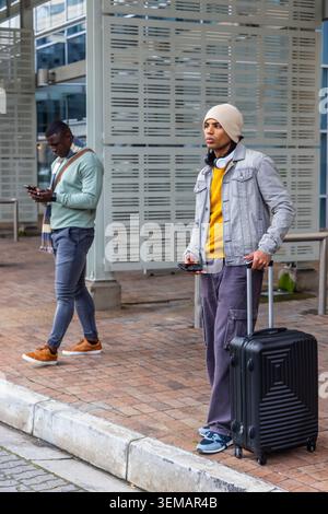 Diversi viaggiatori di sesso maschile in piedi sul marciapiede con valigia nera, berretto e telefoni Foto Stock