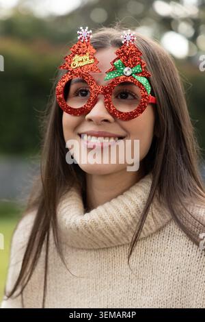 Donna sorridente indossando occhiali rossi sull'albero, fiocco verde a pois e maglione beige in giardino Foto Stock