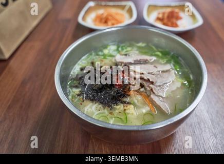 Piatto tradizionale dell'isola di Jeju con filamenti di grano in ricco brodo osseo, condito con carne di maiale bollita a fette, alghe marine e spezie in un bacino metallico rotondo. Foto Stock
