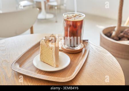 Birra scura ghiacciata e una fetta di crema di dessert in spugna serviti su un vassoio di legno su un tavolo rotondo in un luminoso e moderno Cafe interno. Foto Stock