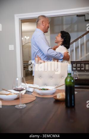 Uomo maturo asiatico e donna anziana asiatica che abbraccia, sorride nella sala da pranzo vicino a un tavolo di legno, vino Foto Stock