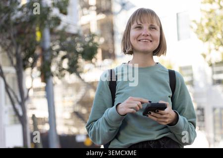 Donna di 20 anni che indossa una felpa verde, uno zaino nero, tiene il telefono, sorride sul marciapiede della città Foto Stock