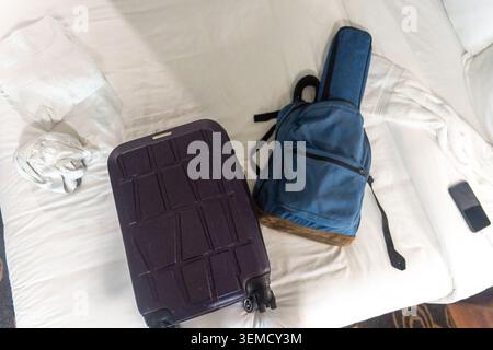 Una valigia rigida viola con ruote si trova su un letto bianco accanto a uno zaino blu. Uno smartphone si trova nelle vicinanze sul materasso, con cuscini e un cr Foto Stock