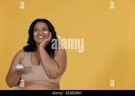 Donna in piedi sullo sfondo della senape che indossa la parte superiore del raccolto beige che regge il vaso di panna che tocca le guance Foto Stock