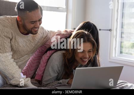 Famiglia di tre persone sdraiata sul letto, papà in maglione crema, bambino in felpa con cappuccio rosa che guarda il laptop Foto Stock