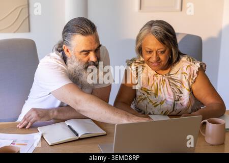 Coppia senior appoggiata a un tavolo di legno, che punta sullo schermo del notebook con carte, tazza, piano floreale Foto Stock