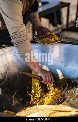 Primo piano di un cuoco che mescola il tradizionale plov uzbeko in un grande calderone kazano, che serve riso fragrante con carne e carote in un mercato all'aperto dell'Uzbe Foto Stock