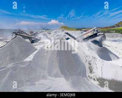 Un'operazione in cava di granito mostra le macchine che lavorano per frantumare e lavorare la pietra. Grandi pile di granito grigio e bianco si trovano sullo sfondo sotto Clear Foto Stock