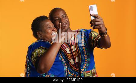 Coppia etnica sorridente che scatta foto con il telefono in studio, divertendosi a fare ricordi con la fotocamera dello smartphone. Le persone allegre scattano foto mentre indossano abiti tradizionali. Foto Stock