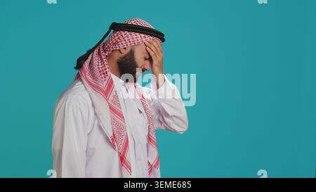 Un tizio del Medio Oriente che ha un terribile mal di testa e tensione, soffre di emicrania su sfondo blu. Giovane uomo che indossa il costume nazionale arabo, si sente malato e stressato con disagio sinusale. Foto Stock