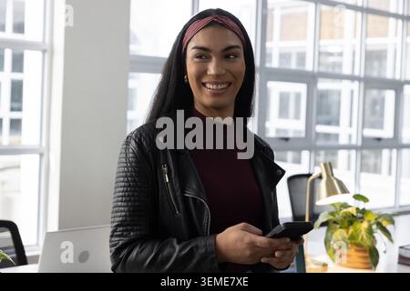 Lavoratrice adulta in piedi vicino alla scrivania in un ufficio moderno, con giacca trapuntata e smartphone in mano Foto Stock