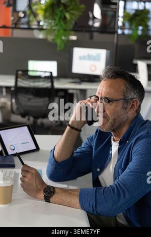 Uomo di mezza età che tiene in mano smartphone e penna, esamina le carte sul tablet con una tazza di caffè, copia spazio Foto Stock