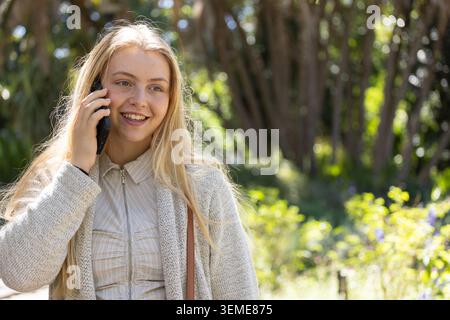 Donna che tiene lo smartphone all'orecchio, sorride e in piedi in un parco e indossa un cardigan e una borsa a spalla Foto Stock