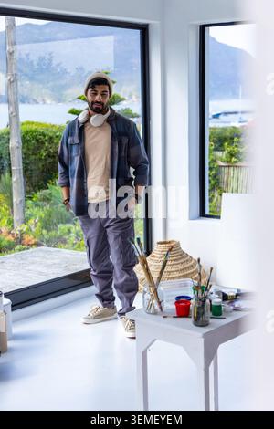 Uomo degli anni '30 in piedi davanti a una finestra in uno studio di casa, con berretto e cuffie che sistemano pennelli Foto Stock