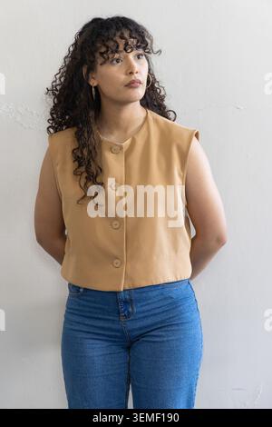 Donna di 20 anni in piedi contro una parete testurizzata, con top senza maniche abbronzatura, jeans blu, cerchi dorati Foto Stock
