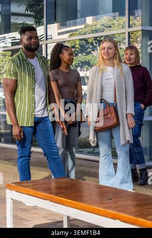 Gruppo di quattro amici che camminano e chiacchierano sul passaggio pavimentato da finestre di vetro e panchina di legno Foto Stock
