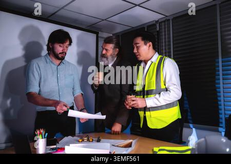 Tre persone esaminano i progetti a tarda notte, discutendo dei piani. Lavorano nell'organizzazione di riunioni di gruppo, nella revisione del progetto di progettazione con l'azienda senior c Foto Stock