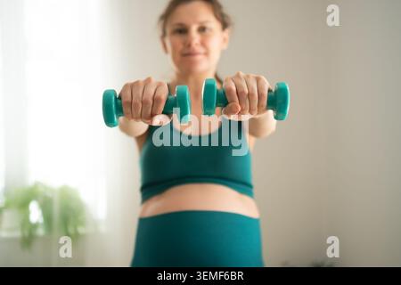 Donna incinta braccia allungato tenendo i manubri per l'allenamento di forza a casa. Ti aspetti che la madre dia un tono al corpo attraverso l'allenamento di resistenza con pesi leggeri. La futura mamma rimane potente mantenendo la forma fisica Foto Stock