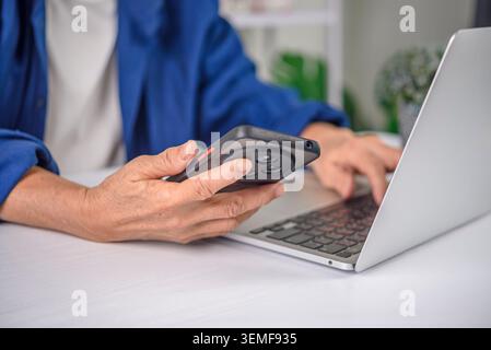 Primo piano di una donna anziana che tiene le mani sullo smartphone e digita sulla tastiera del laptop. Donna d'affari anziana che lavora in multitasking con tecnologia, immissione dati e d Foto Stock