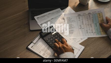 Persona che calcola i moduli fiscali con calcolatore e documenti finanziari sulla scrivania, supportando il flusso di lavoro di preparazione fiscale, la revisione delle spese, il reddito Foto Stock