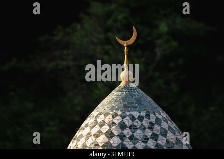 La mezzaluna dorata risplende in cima alla struttura a cupola. Le piastrelle a motivi geometrici riflettono la luce soffusa della sera. Il fogliame verde scuro incornicia la scena. La quiete serena suggerisce una tranquilla riverenza. Ideale per spirituali, culturali Foto Stock