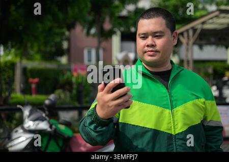 Un ritratto orizzontale professionale di un corriere o autista thailandese maggiorenne con un dispositivo smartphone nero. Il soggetto è vestito di ciao Foto Stock