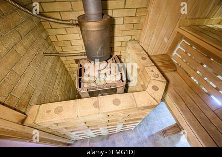 accogliente sauna interna con panche in legno, accenti in mattoni e una stufa in metallo con rocce Foto Stock