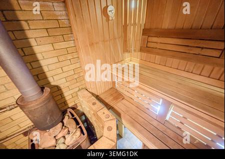 accogliente sauna interna con panche in legno, accenti in mattoni e una stufa in metallo con rocce Foto Stock