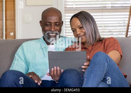 Coppia matura che condivide il tablet sul divano grigio di casa, indossando camicie color tè e ruggine, sorridendo Foto Stock