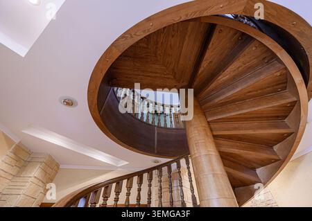 la foto dall'angolo basso presenta una scala a spirale in legno, elegantemente curvilinea verso l'alto verso un soffitto bianco con illuminazione a incasso. Foto Stock