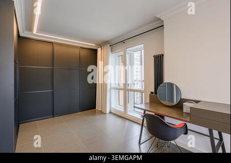 Camera minimalista con armadio scuro, scrivania con specchio rotondo e grandi finestre. Pavimento piastrellato, tonalità neutre e un'estetica moderna. Foto Stock