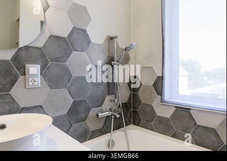 Bagno moderno con piastrelle esagonali grigie e bianche, lavandino e vasca bianchi e doccia cromata. La luce naturale riempie lo spazio Foto Stock