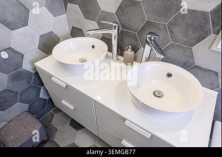 Bagno moderno con due lavandini bianchi, rubinetti cromati e piastrelle geometriche grigio/bianco. Design pulito ed elegante. Foto Stock