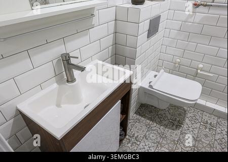 il bagno moderno presenta piastrelle bianche della metropolitana, un elegante lavandino bianco con rubinetto d'argento montato su un mobile in legno e un wc a parete su patte Foto Stock