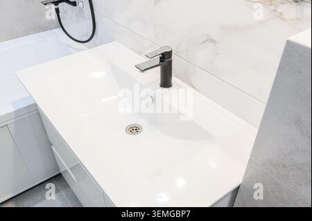 il bagno era pulito e aveva un lavandino rettangolare bianco con un moderno rubinetto nero. Lo sfondo include una vasca da bagno bianca e piastrelle con motivi in marmo Foto Stock
