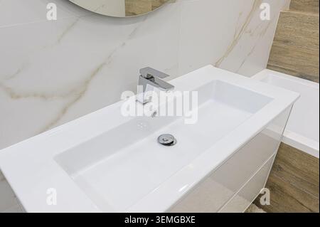 primo piano di un moderno lavandino del bagno con rubinetto cromato. Il lavandino è bianco e rettangolare, adagiato su piastrelle in marmo e legno Foto Stock