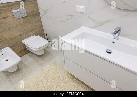 bagno luminoso con lavandino bianco, wc e bidet. Le pareti sono piastrellate con motivi in marmo e legno. Un soffice tappeto si trova sul til Foto Stock