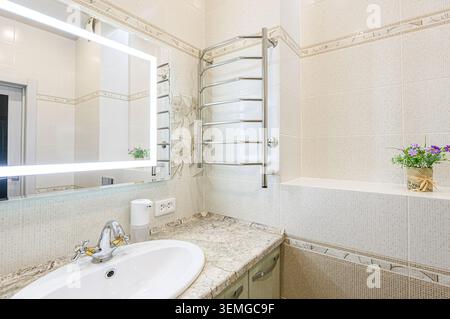 bagno ben illuminato con piastrelle beige, un moderno specchio illuminato, un lavandino con ripiano in granito e uno scalda asciugamani. Una pianta decorativa in vaso aggiunge Foto Stock