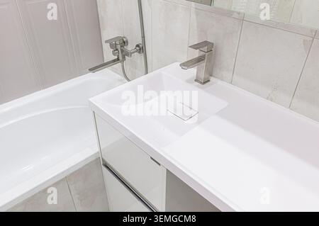 Bagno luminoso con lavandino bianco, rubinetto moderno, vasca con doccia e piastrelle neutre Foto Stock
