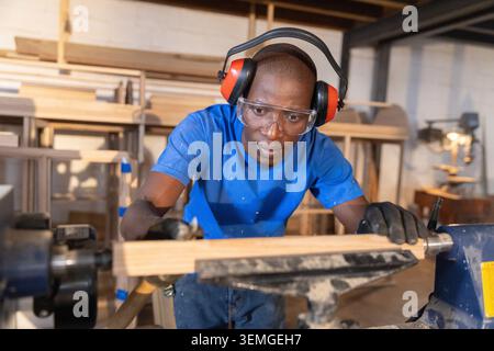 Falegname afro-americano di medie dimensioni maschio che modellano il raggio sul tornio indossando guanti da lavoro in officina Foto Stock