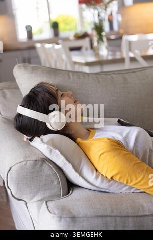 Donna asiatica sdraiata sul divano, con cuffie bianche, smartphone appoggiato sul petto vicino alla cucina Foto Stock