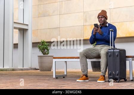 Uomo seduto che controlla il telefono sulla panchina del plaza con berretto marrone, valigia e cuffie Foto Stock