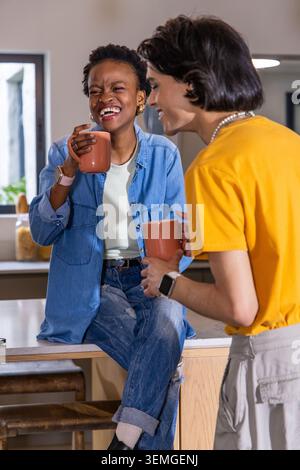 Coppie diverse che tengono tazze abbinate, ridono e si appoggiano in cucina, jeans e camicie senape Foto Stock