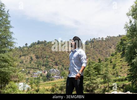 Un colpo ad angolo basso di un giovane Sikh che indossa un turbante nero e abiti formali che guarda verso il cielo mentre posa a Himachal Pradesh, India. Foto Stock
