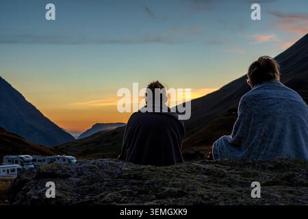 Due donne da dietro, avvolte da calde coperte, siedono su un grande masso contemplando il sole tramontare dietro le montagne. Una suggestiva luce dorata Foto Stock