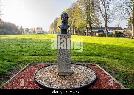 Un monumento commemorativo con un busto in bronzo scuro sorge su un piedistallo in pietra all'interno di un'aiuola circolare. Sullo sfondo si estende un vasto prato verde Foto Stock
