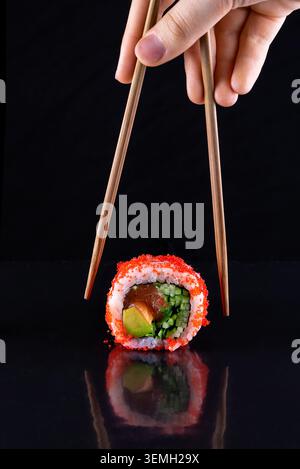 Le bacchette raccolgono un grande rotolo di sushi futomaki multi ingrediente su una superficie scura riflettente Foto Stock